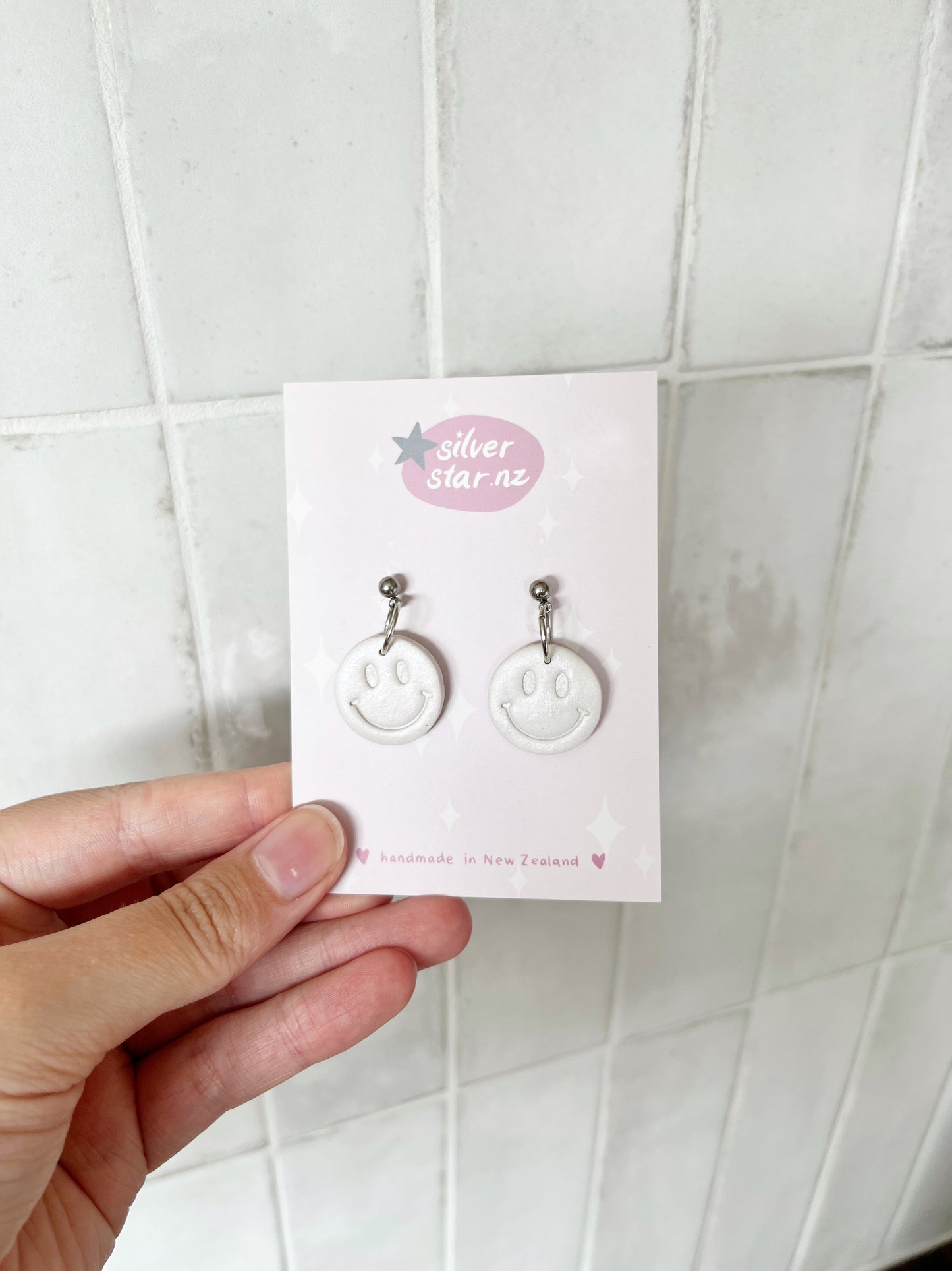 A hand holds a card featuring Smiley Polymer Clay Earrings—white, round handmade earrings with smiley faces and stainless steel hooks by silverstar.nz—set against a tiled white background.