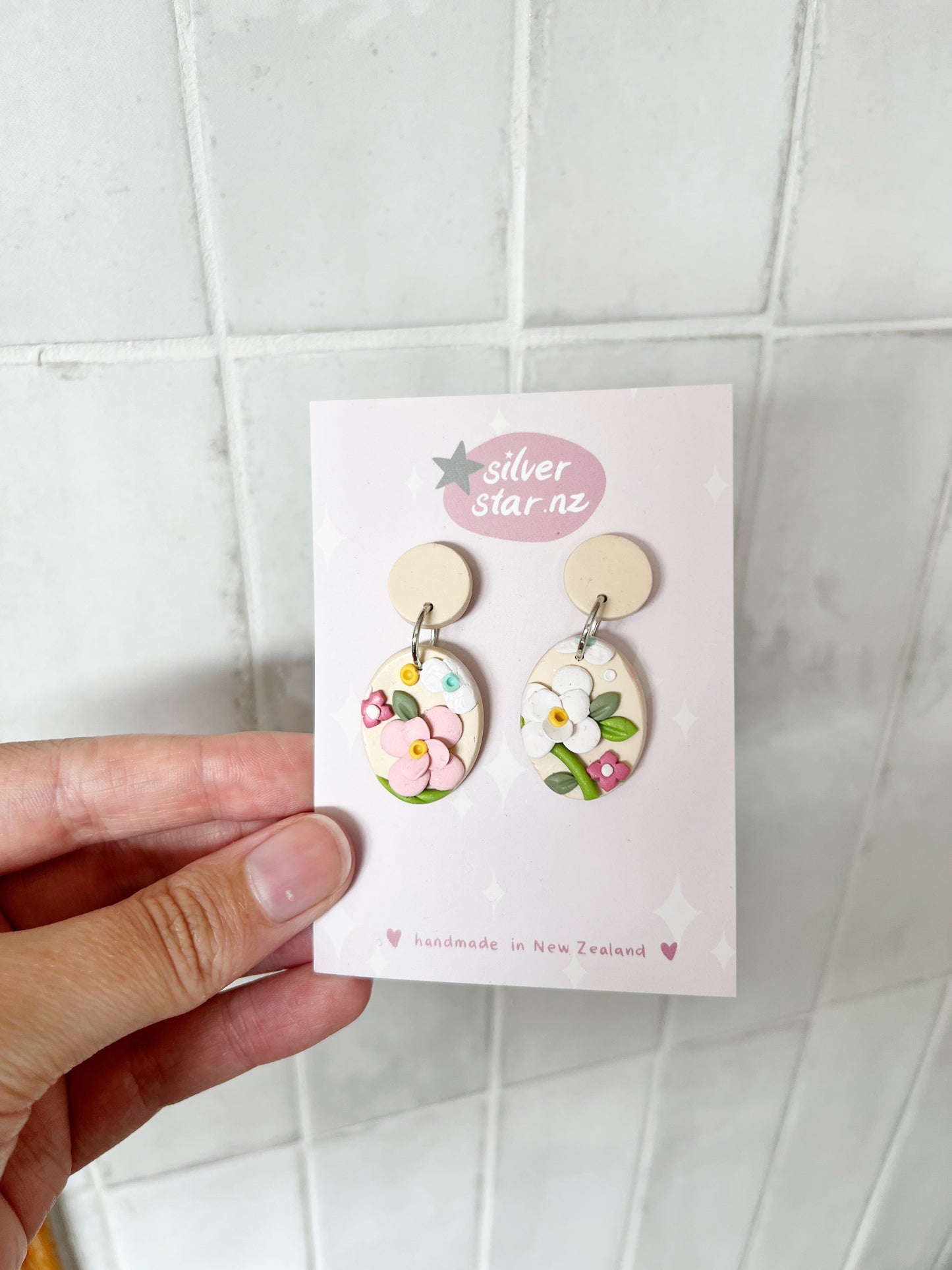 A hand holds a card displaying Floral Organic Oval Polymer Clay Earrings with pink, yellow, and white floral designs. The earrings have stainless steel hooks. The card reads silverstar.nz and handmade in New Zealand against a tiled background.