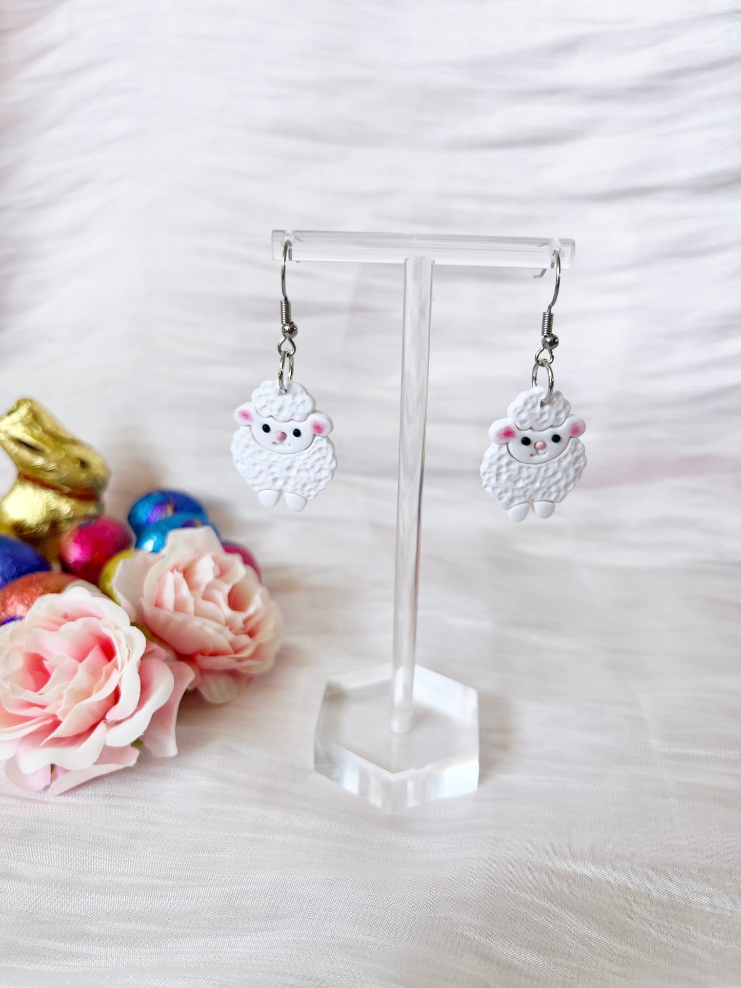 The Fluffy Sheep Polymer Clay Earrings by silverstar.nz are showcased on a clear stand, featuring charming pink accents. Pink roses and colorful metallic eggs in the background enhance the whimsical appeal of these unique handmade earrings.