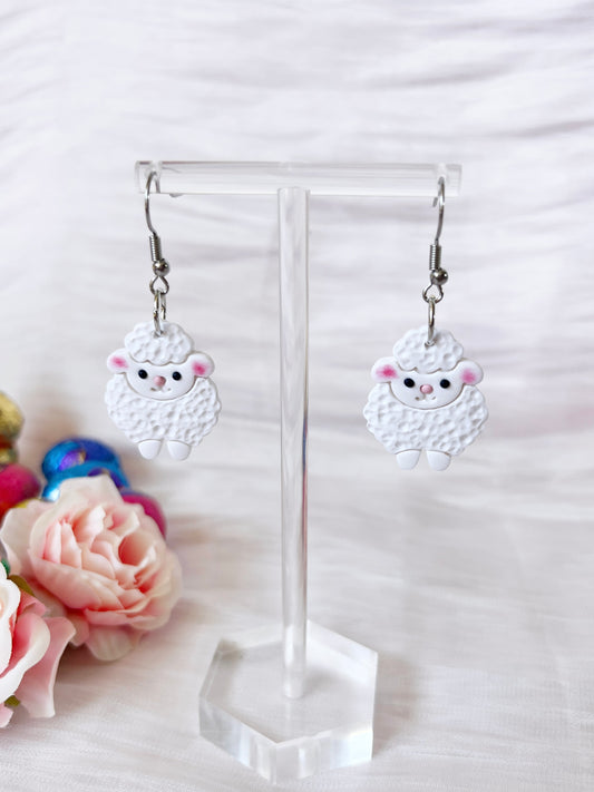 Fluffy Sheep Polymer Clay Earrings by silverstar.nz dangle on a transparent stand, featuring sheep with pink cheeks and black eyes. Pink roses and colorful wool balls enhance the unique design, all set against a soft white fabric backdrop.