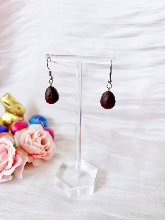 Mini Creme Egg Polymer Clay Earrings by silverstar.nz hang elegantly on a transparent stand. The backdrop features decorative pink roses, colorful chocolate eggs, and a golden chocolate bunny on the left, highlighting these stunning handmade earrings in a bright and elegant setting.
