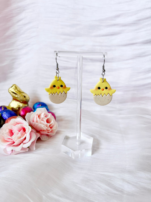 Egg Baby Chick Polymer Clay Earrings by silverstar.nz feature yellow chicks hatching from eggs on a clear stand, highlighted by a gold bunny and colorful eggs, with pink roses on a white textured surface in the background.