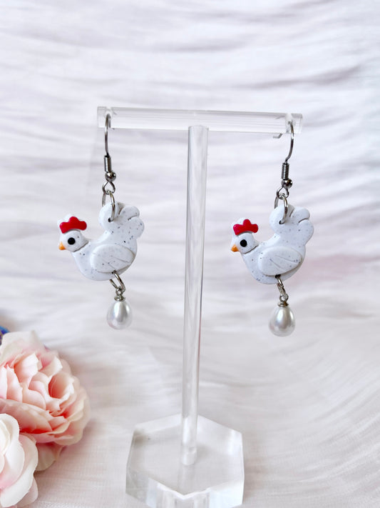 Chicken with Egg Polymer Clay Earrings by silverstar.nz feature handmade white chicken charms with red combs and dangling faux pearl beads, shown on a clear stand with a soft pink background and a nearby pink flower.