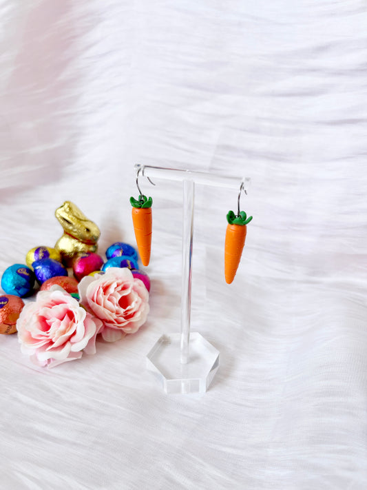 The Carrot Polymer Clay Earrings by silverstar.nz are showcased on a clear stand, surrounded by pink roses, a golden bunny, and colorful Easter eggs against a soft white background.