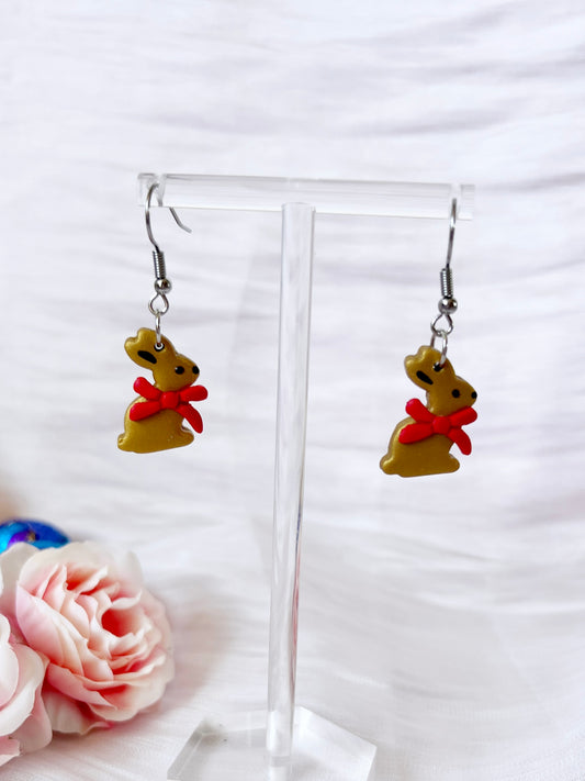 Gold Bunny Polymer Clay Earrings by silverstar.nz feature handmade brown bunnies with red bows, shown on a transparent stand with soft pink flowers in the background, highlighting their funky and charming design.