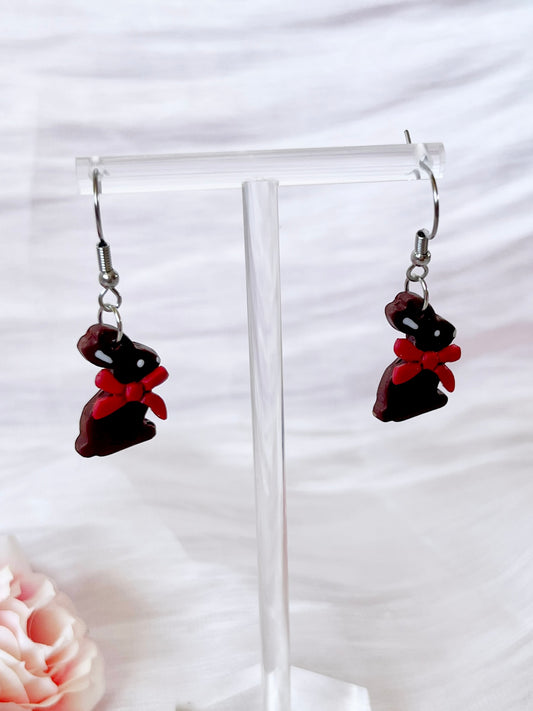 The Chocolate Bunny Polymer Clay Earrings by silverstar.nz feature bunny charms with black bodies and red bows. Displayed on a clear stand, these handmade earrings have a soft light backdrop with a hint of pink flower in the corner.