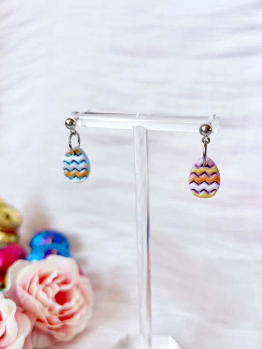 Two handmade Decorated Eggs Polymer Clay Earrings by silverstar.nz, featuring colorful zigzag patterns, hang from a clear stand with stainless steel hooks. Pink roses and blurred multicolored decorations in the background add charm to their unique funky design.