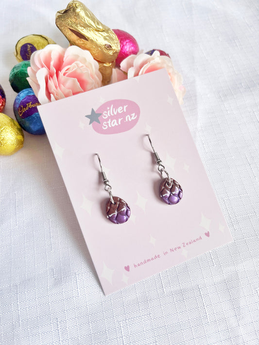 Opened Easter Egg Polymer Clay Earrings by silverstar.nz, featuring a unique purple dragon-scale design, rest on a card. They are surrounded by pink flowers, a gold bunny, and colorful foil-wrapped chocolates on a textured white background.
