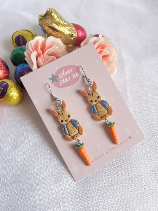 Peter Rabbit Polymer Clay Earrings by silverstar.nz feature a rabbit in a blue jacket holding carrots, crafted from handmade polymer clay. Displayed on a silver star nz card, these earrings have stainless steel hooks and are surrounded by colorful chocolates and flowers for a funky design.