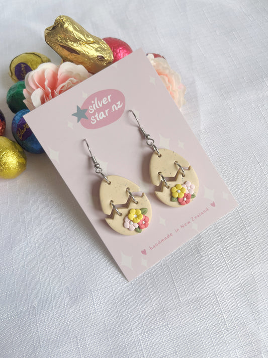 Broken Egg Polymer Clay Earrings from silverstar.nz feature cracked egg shapes with flowers and smiles, crafted using polymer clay and stainless steel hooks. Theyre displayed on a silver star nz card, surrounded by pastel chocolate eggs, a pink flower, and gold-wrapped chocolate on white.