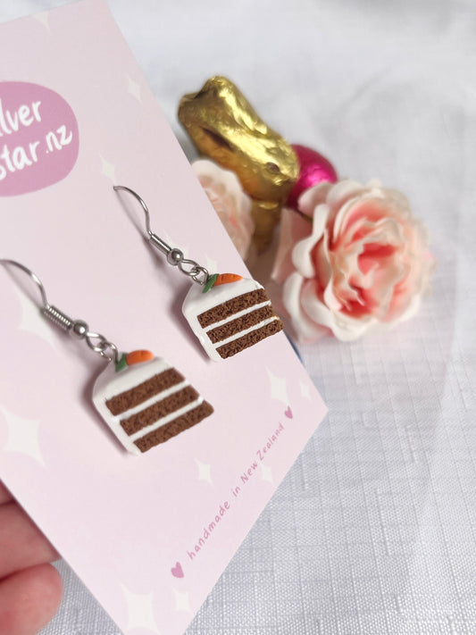 A close-up of the Carrot Cake Polymer Clay Earrings by silverstar.nz highlights a cake slice motif on the card. Set against a pink rose and gold-accented background, these New Zealand handmade earrings feature layered frosting and a carrot topper.
