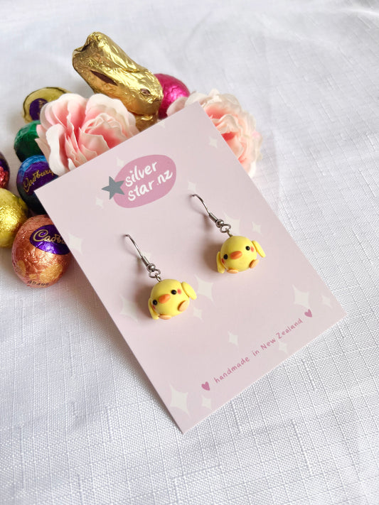 A pair of Mini Baby Chick Polymer Clay Earrings by silverstar.nz are displayed on a card labeled silverstar.nz. The card sits on a white surface surrounded by pink flowers and foil-wrapped chocolate eggs, showcasing their unique design.