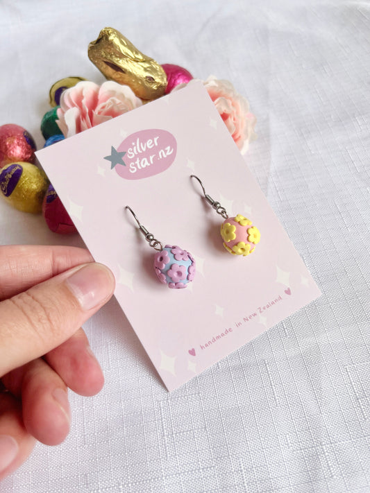 A hand holds Floral Egg Polymer Clay Earrings with stainless steel hooks on a silverstar.nz card. The pink and purple floral earrings are set against colorful Easter eggs and pink roses in the background.