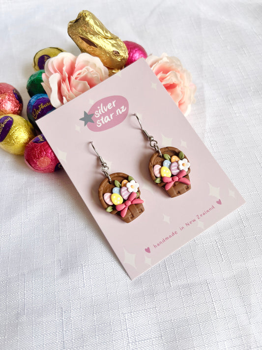 A pair of unique, handmade Egg Basket Polymer Clay Earrings by silverstar.nz features colorful eggs and flowers, presented on a branding card amidst chocolates and rose petals on a white textured surface.