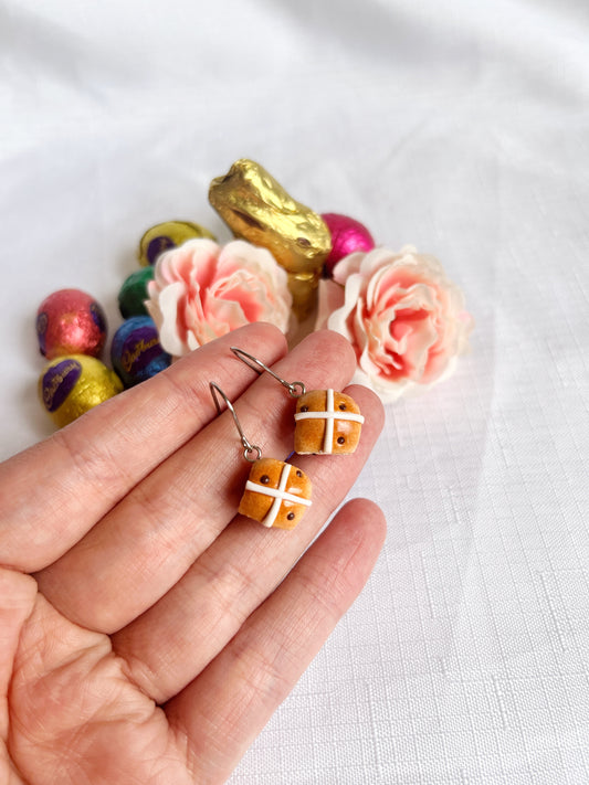 A hand showcases silverstar.nzs Hot Cross Bun Polymer Clay Earrings with pink roses, a gold foil chocolate bunny, and colorful foil-wrapped eggs in the background on a pristine white surface.