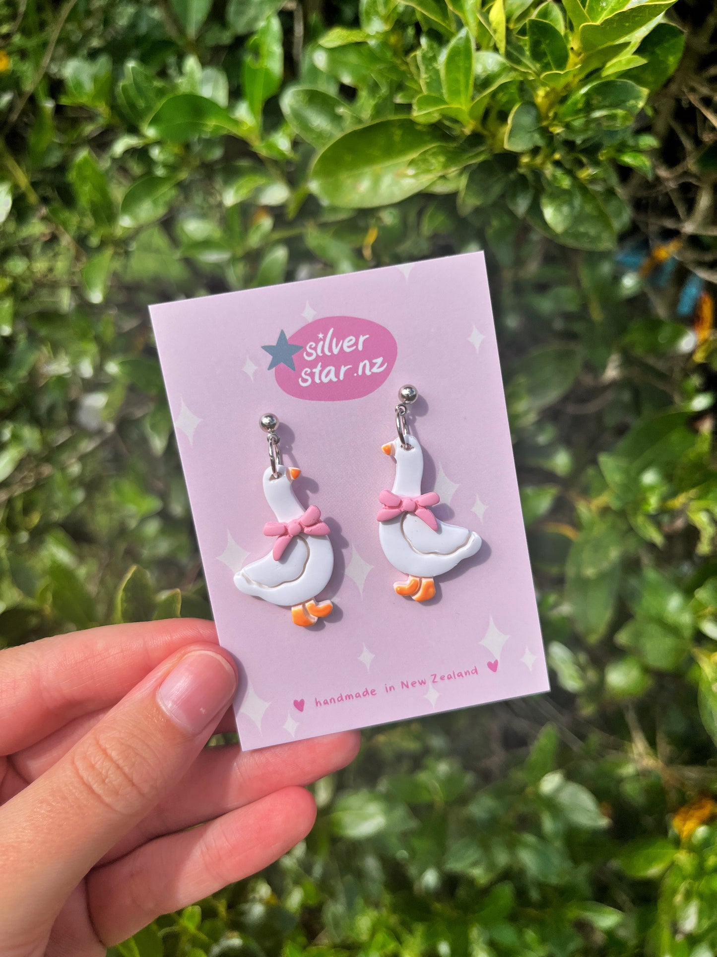A hand holds a card reading silverstar.nz with Goose with Bow Polymer Clay Earrings, handmade in New Zealand, shaped like white geese with pink bows, set against leafy green foliage.