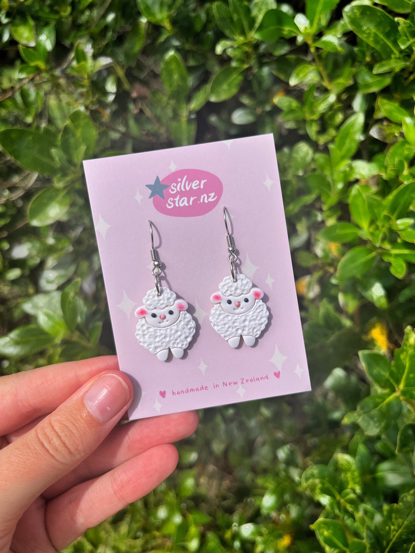 A person holds Fluffy Sheep Polymer Clay Earrings on a light purple card with silver stars, reading silverstar.nz and handmade in New Zealand, set against a green foliage backdrop.