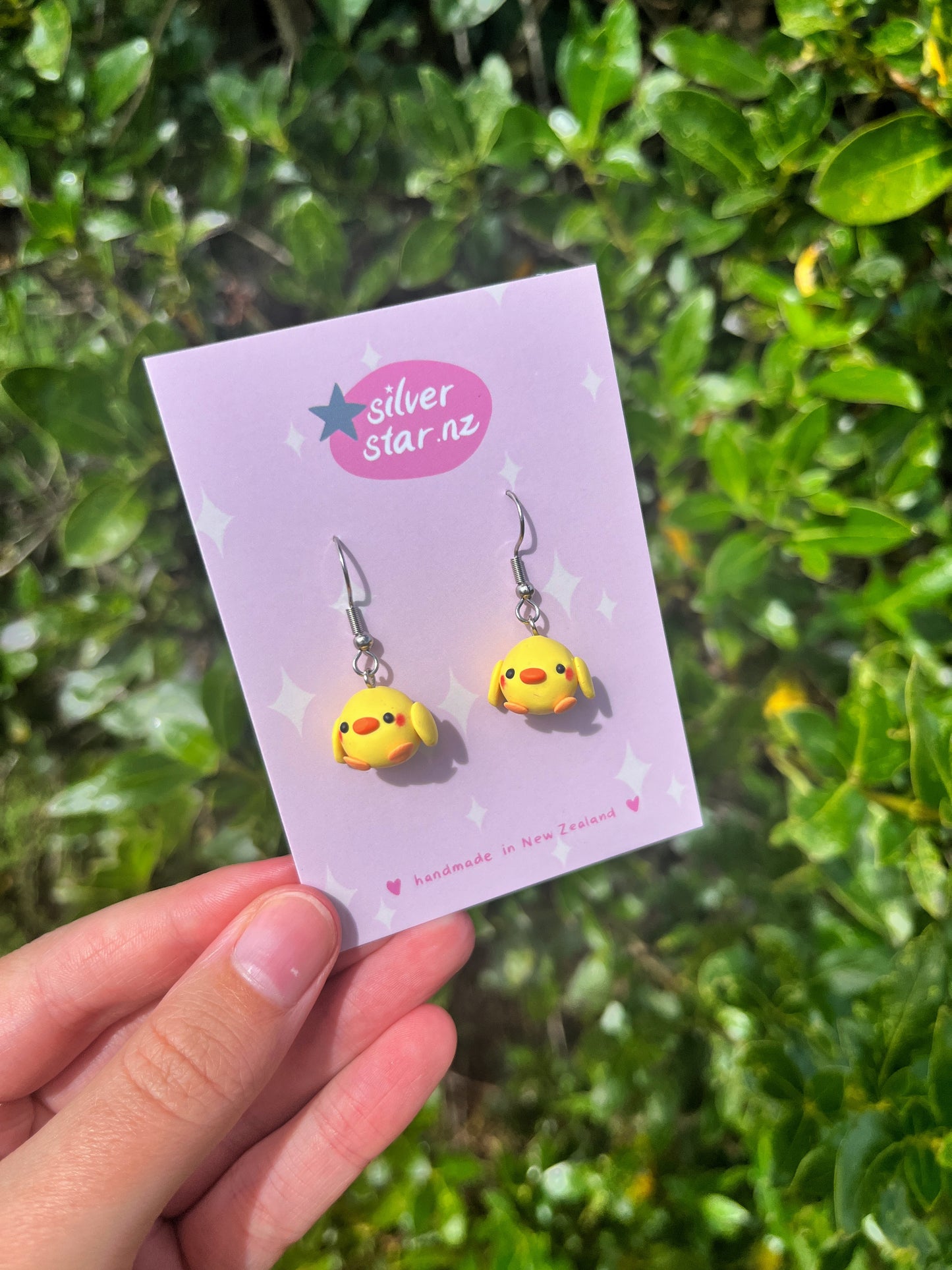 A hand displays silverstar.nz Mini Baby Chick Polymer Clay Earrings with charming yellow and red characters featuring long ears, set against a lush green foliage background, accentuating their playful design.