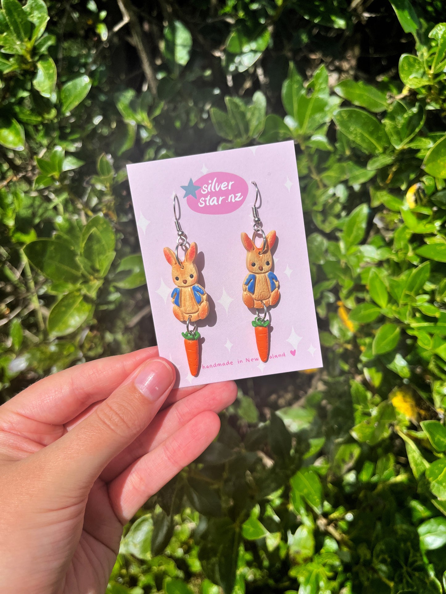 A hand displays Peter Rabbit Polymer Clay Earrings with bunny and carrot designs against green foliage. The card features silverstar.nz with stars and Handmade in New Zealand, highlighting these unique creations.
