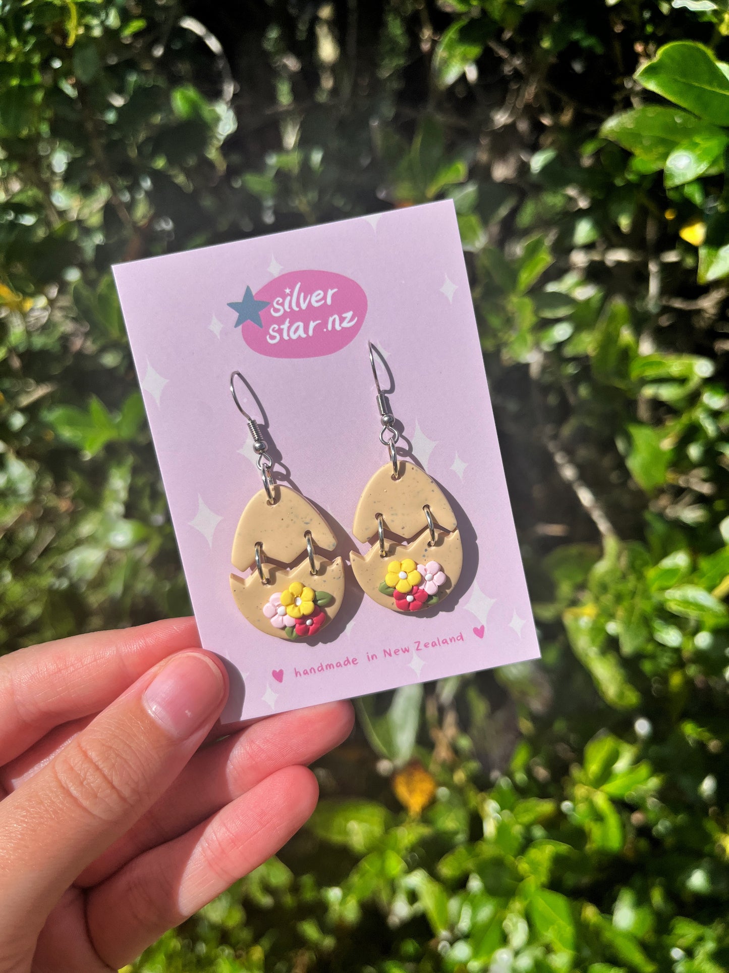A person holds the Broken Egg Polymer Clay Earrings featuring teardrop shapes with colorful flowers on a card labeled silverstar.nz in pink. Green foliage in the sunny background highlights the stainless steel hooks.