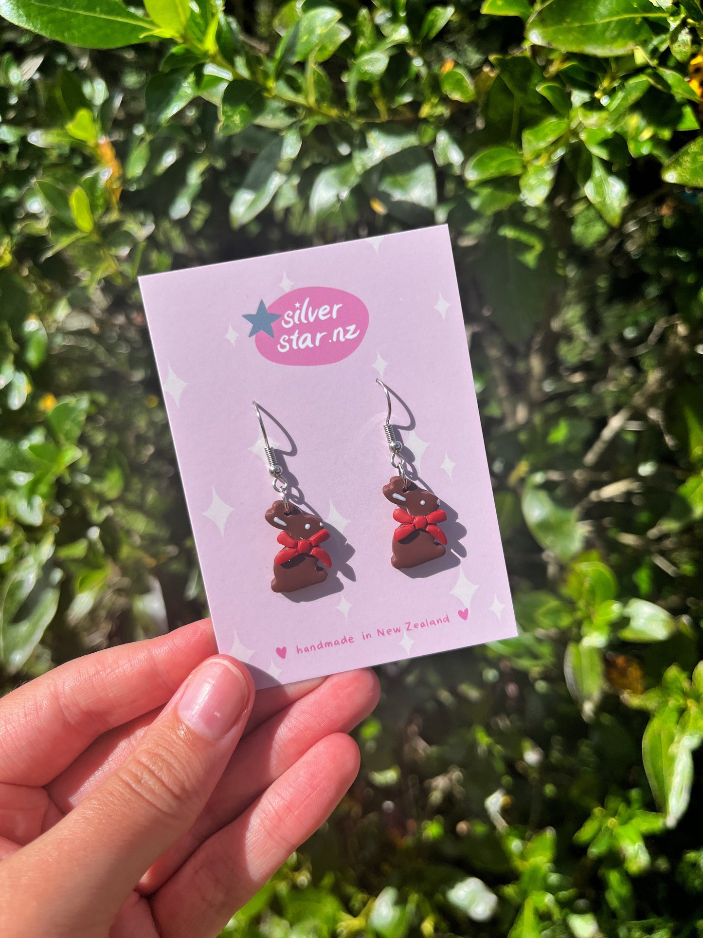 A hand holds a pink card labeled silverstar.nz, which says handmade in New Zealand, displaying Chocolate Bunny Polymer Clay Earrings with funky design elegance against lush green leaves.