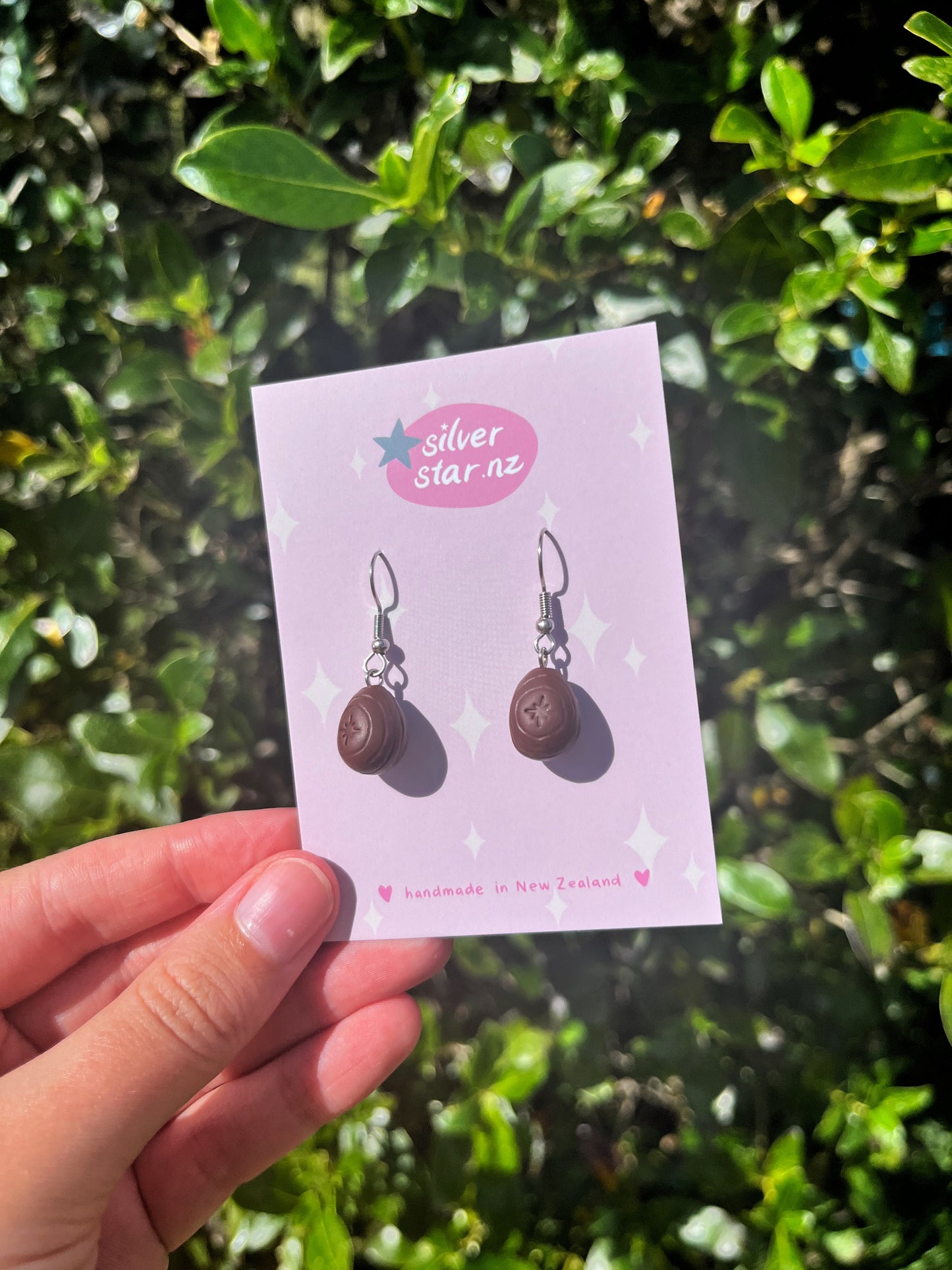A close-up of silver hook Mini Creme Egg Polymer Clay Earrings, resembling chocolates, on a light purple silverstar.nz card stating handmade in New Zealand, set against lush green foliage.