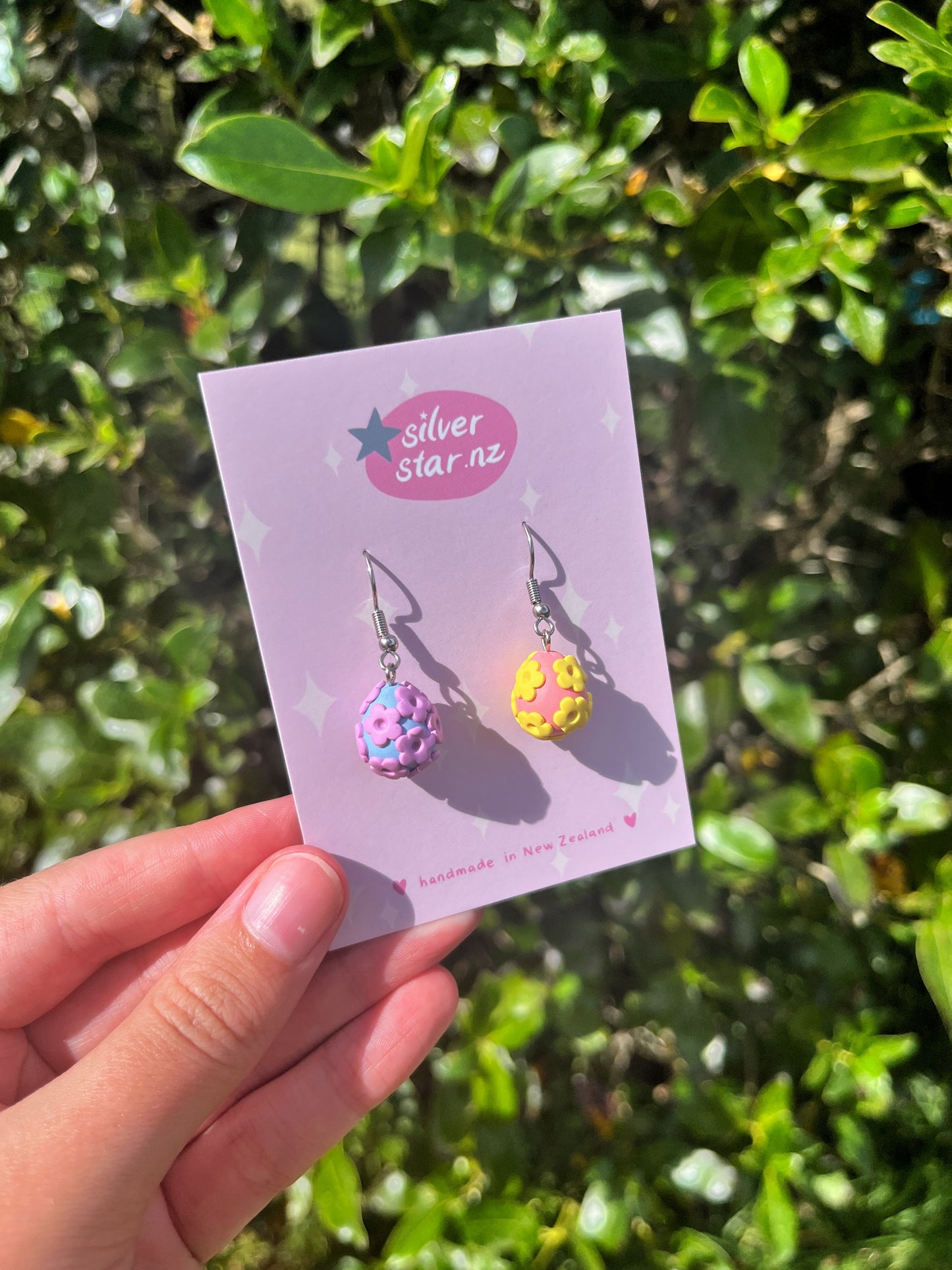 A hand displays the Floral Egg Polymer Clay Earrings by silverstar.nz on a lavender card. One earring is pink with blue sprinkles and the other is yellow with purple, both creating a vivid contrast against lush green foliage.