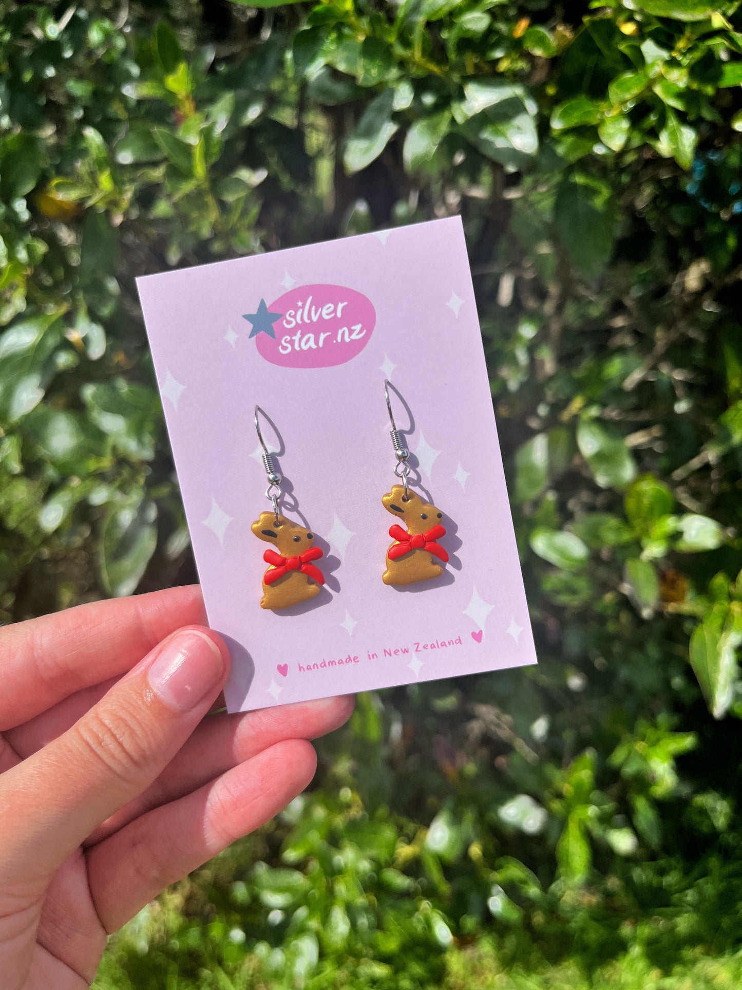 A hand holds a silverstar.nz card showing handmade Gold Bunny Polymer Clay Earrings with stainless steel hooks. The card states theyre handmade in New Zealand, with lush green foliage softly blurred in the background.