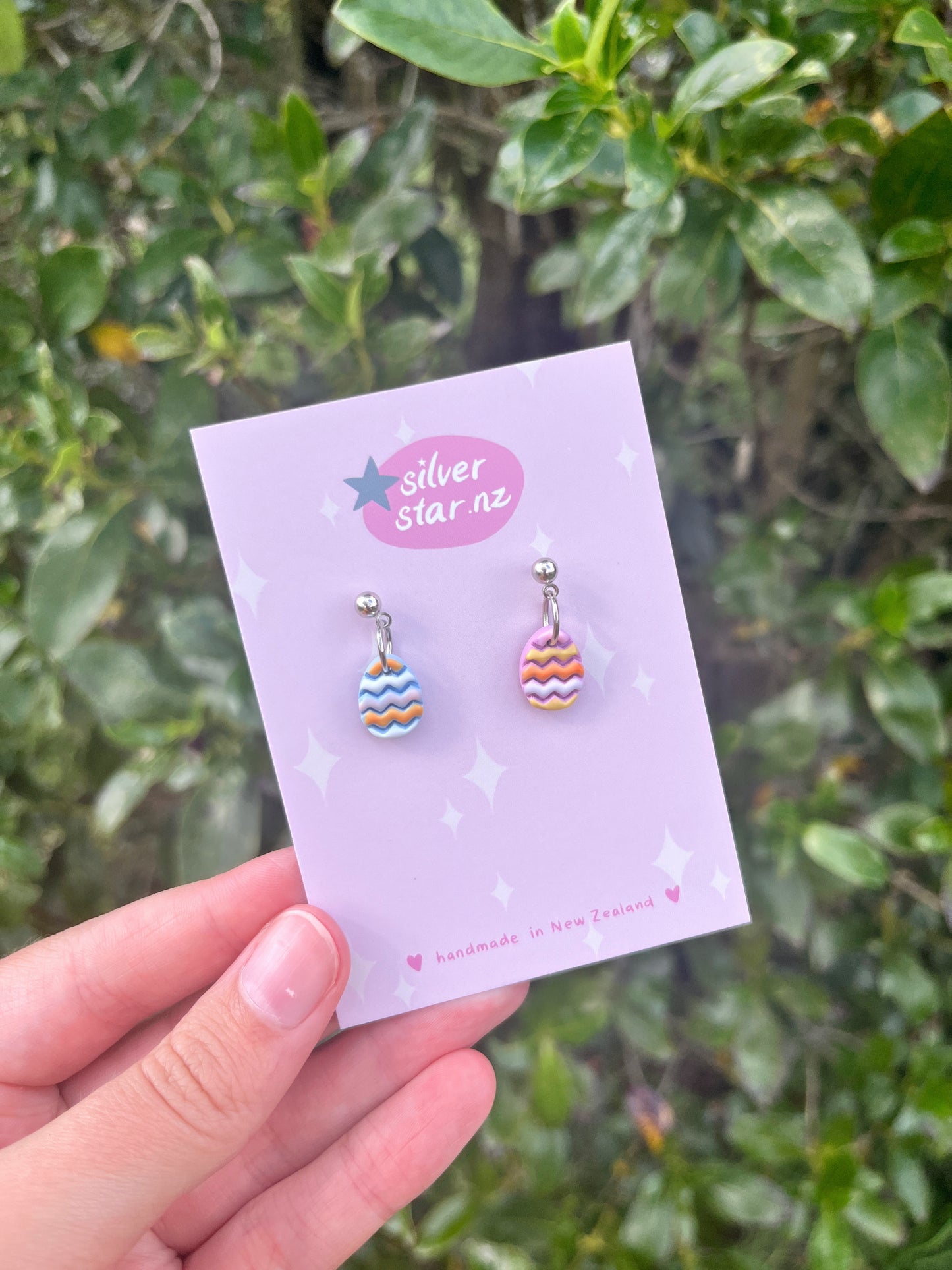 A hand holds a card against green foliage, displaying Decorated Eggs Polymer Clay Earrings with colorful wavy patterns and stainless steel hooks. The silverstar.nz logo features a star and notes handmade in New Zealand, emphasizing the unique funky design of these earrings.