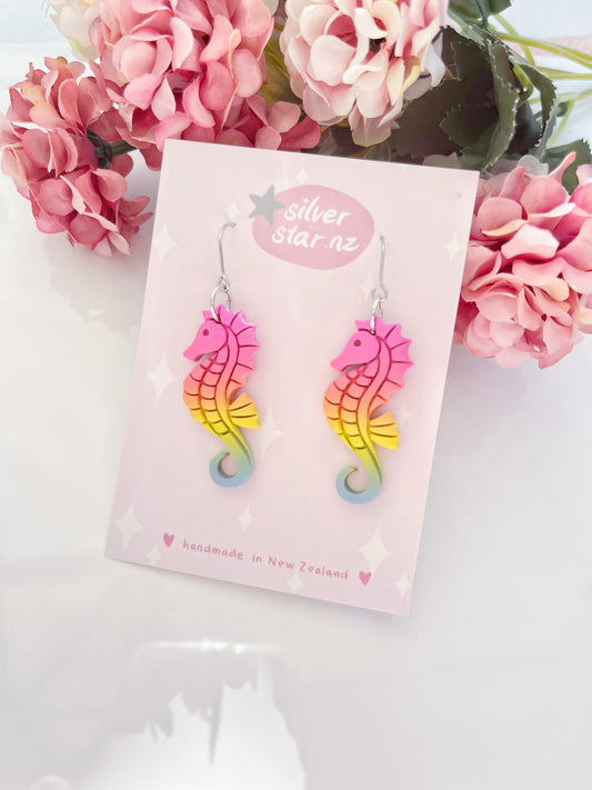 The Rainbow Seahorse Polymer Clay Earrings, featuring a vibrant blend of pink, yellow, and green, are presented on a card adorned with the silverstar.nz logo. These handmade earrings, designed in New Zealand with unique artistry, are accompanied by an array of pink and white flowers.