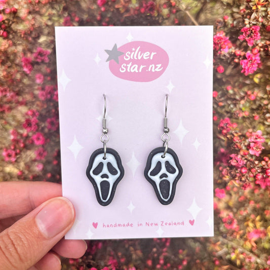 The Ghostface Polymer Clay Earrings dangle from a display card that features "silverstar.nz" and "handmade in New Zealand." A hand showcases the card against a backdrop of small pink flowers, emphasizing their distinctive design.