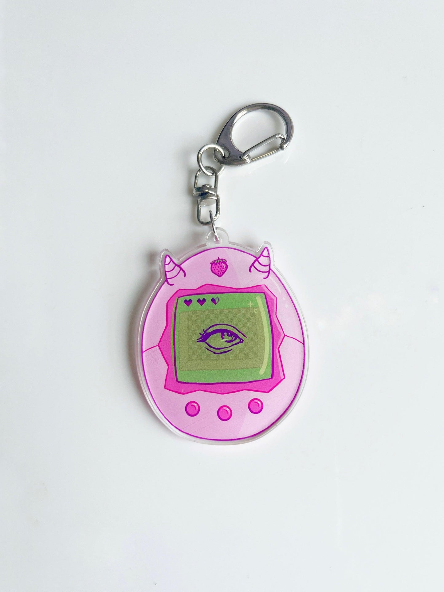The Strawberry Tamagotchi Acrylic Keyring is an artist-designed pink acrylic keychain shaped like a retro virtual pet with two small horns. Its screen features a green eye, enhancing its whimsical charm. The keyring includes a metal ring at the top and is presented on a white surface.