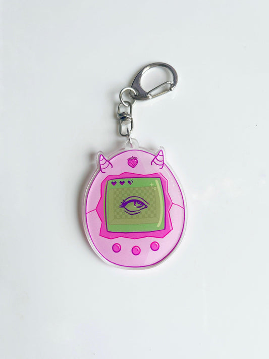 The Strawberry Tamagotchi Acrylic Keyring is an artist-designed pink acrylic keychain shaped like a retro virtual pet with two small horns. Its screen features a green eye, enhancing its whimsical charm. The keyring includes a metal ring at the top and is presented on a white surface.