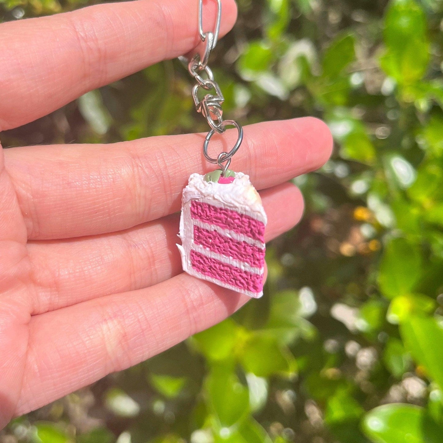 A hand holds the Strawberry Cake Polymer Clay Keyring, featuring a miniature slice of pink and white layered cake with frosting. Its unique, funky design pops against the softly-focused backdrop of green plants.