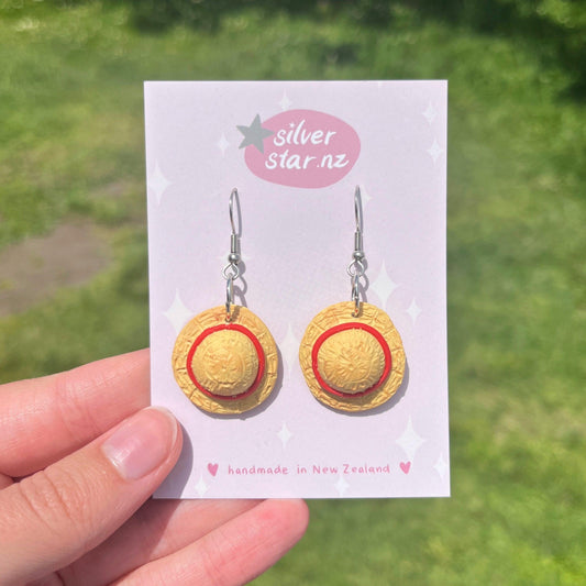 A pair of Luffy Hat One Piece Polymer Clay Earrings, designed to resemble straw hats with a red band, are displayed on a card labeled silver star.nz against a blurred green grassy background, highlighting their unique design.