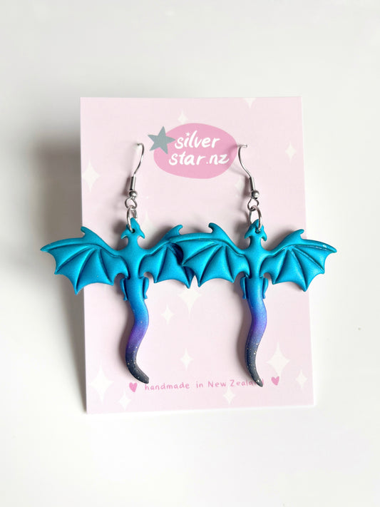The blue and purple dragon-shaped earrings, made from polymer clay, are displayed on a silverstar.nz card adorned with pink and white hues, star designs, and handmade in New Zealand at the bottom, reflecting their funky style.