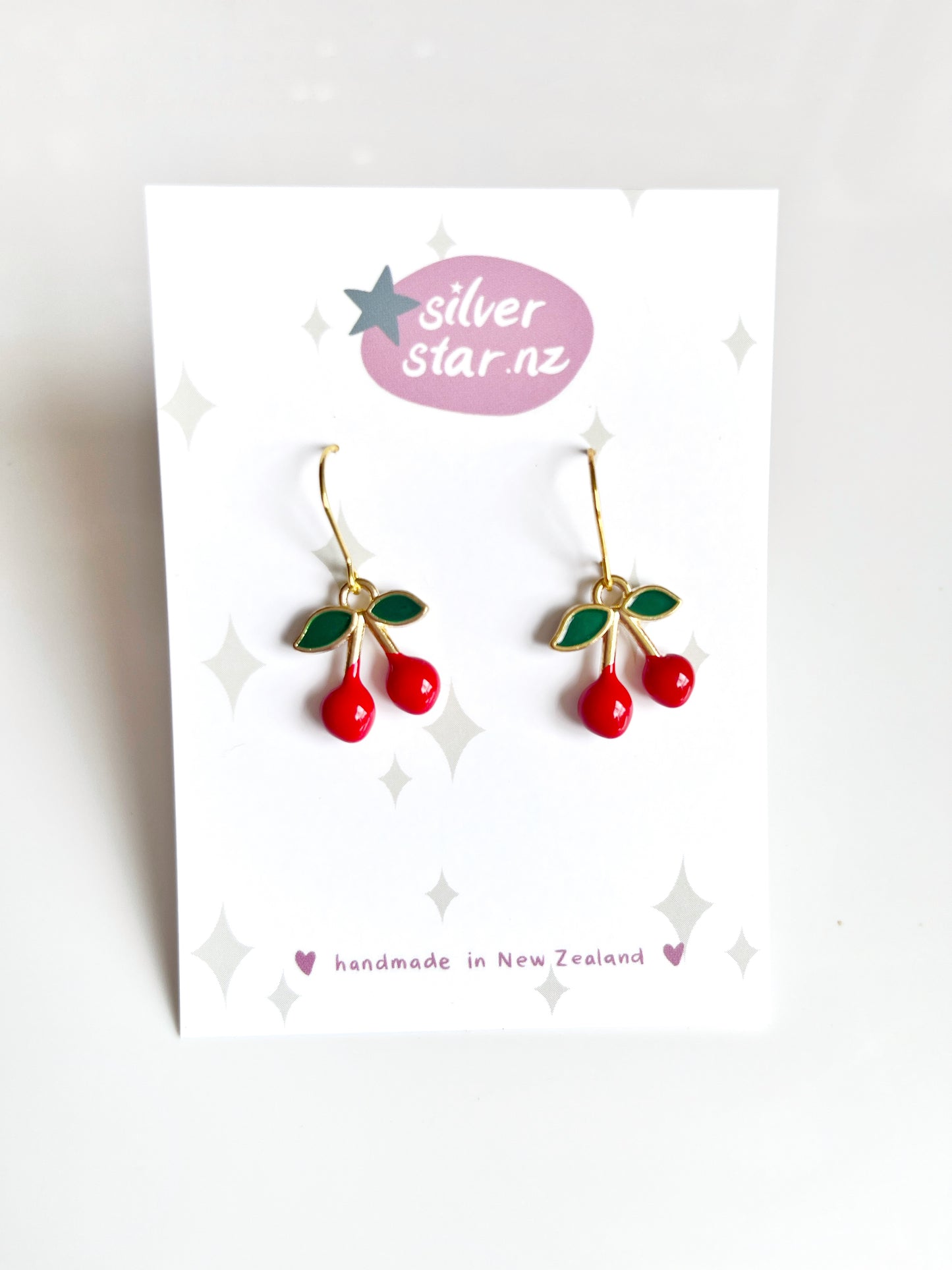 The Mini Cherry Enamel Earrings feature red cherries and green leaves on gold hooks, displayed on a white card. The card includes silverstar.nzs purple logo and notes theyre handmade in New Zealand with durable stainless steel hooks.