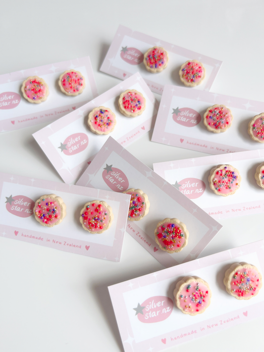 The Hundreds and Thousands Biscuit Stud Earrings by silverstar.nz feature cookie-shaped polymer clay studs with pink icing and colorful sprinkles, displayed on silverstar.nz and handmade in New Zealand cards, artfully scattered on a white surface.
