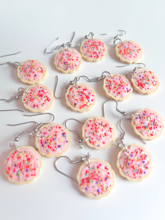 Numerous pairs of silverstar.nzs Hundreds and Thousands Biscuit Earrings, crafted from polymer clay with pink icing, colorful sprinkles, and stainless steel hooks, are displayed on a white surface.