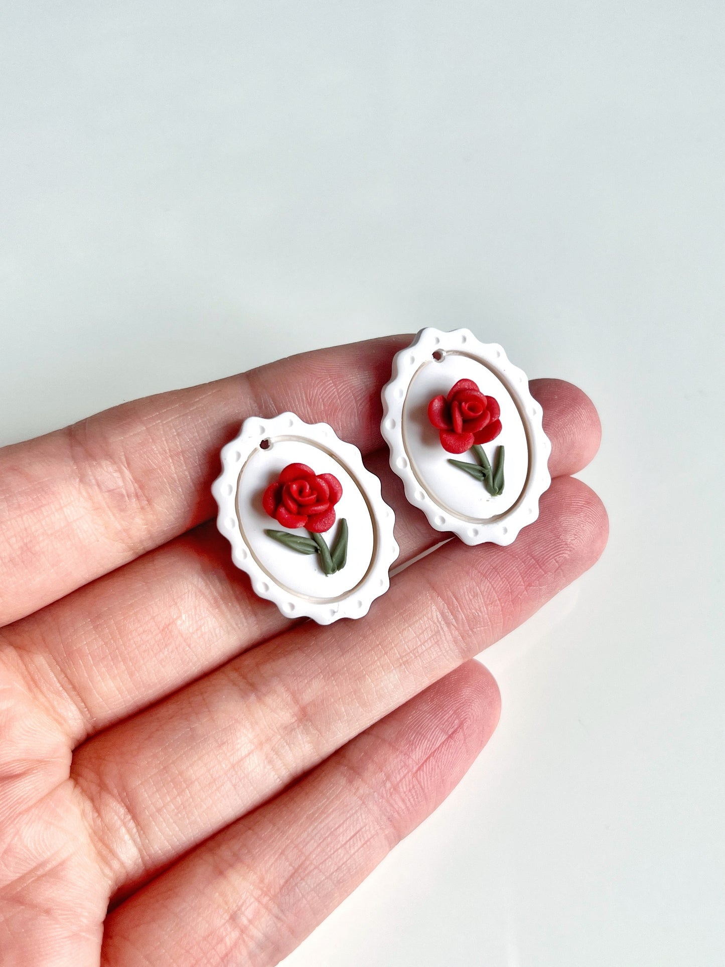A hand displays the Framed Rose Polymer Clay Earrings by silverstar.nz—two funky handmade earrings featuring white scalloped edges, red rose designs, on stainless steel hooks. Theyre set against a plain, light-colored background.