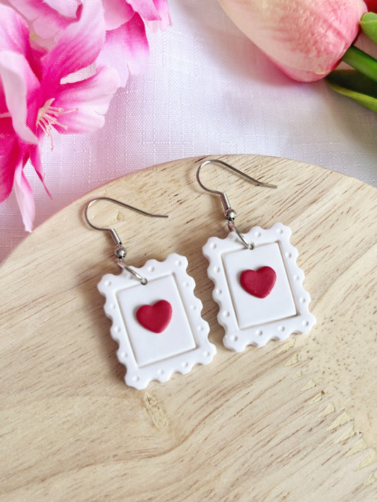 Heart Stamp Polymer Clay Earrings by silverstar.nz feature white picture frame shapes with red hearts in the center, displayed on wood and complemented by pink flowers. Crafted from polymer clay with stainless steel hooks, these handmade earrings are a unique accessory.