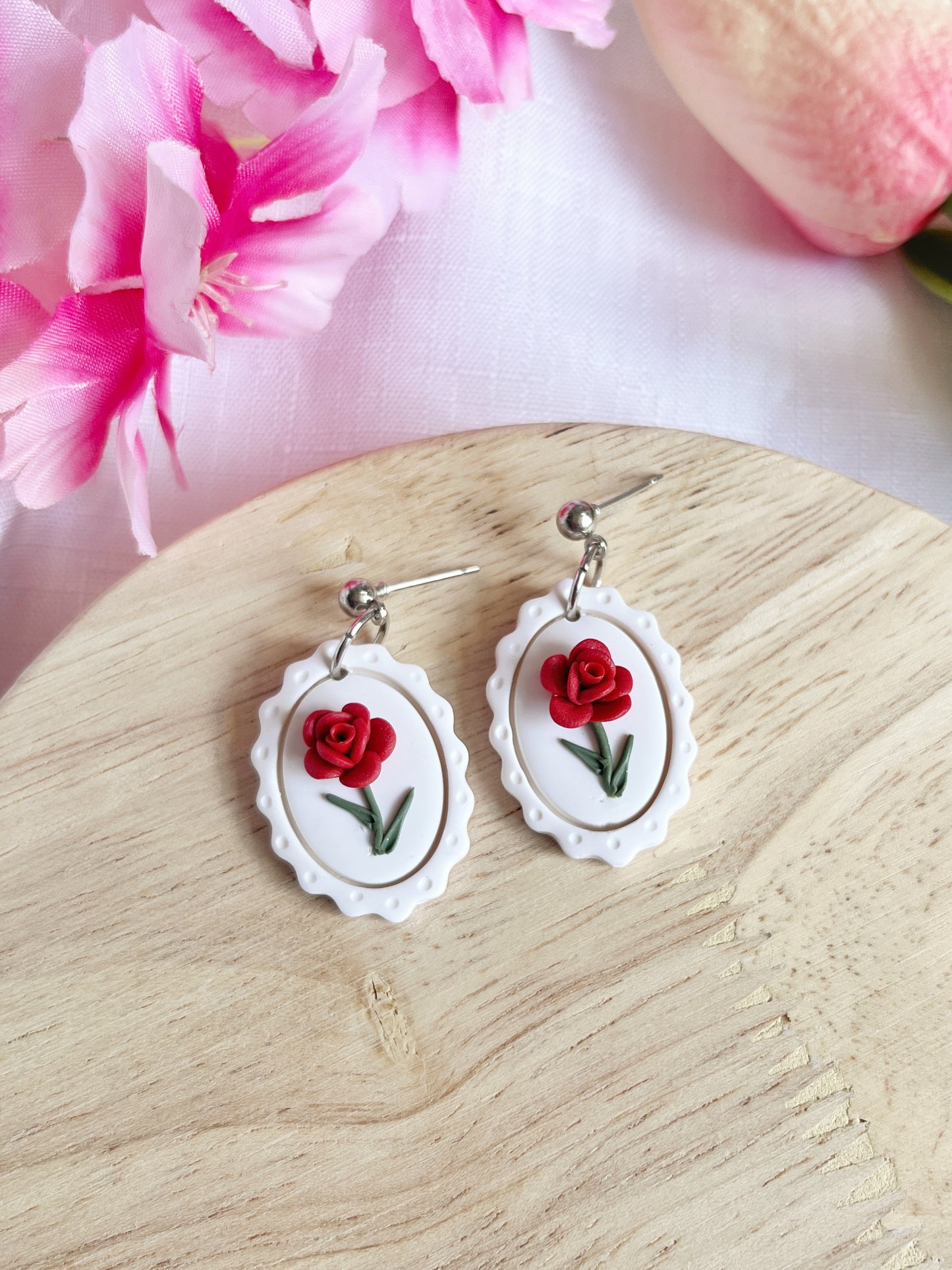 The Framed Rose Polymer Clay Earrings by silverstar.nz, featuring red rose designs on white frames with stainless steel hooks, rest on a wooden surface. Surrounded by pink flowers and a tulip, they add a colorful touch with their unique funky handmade design.