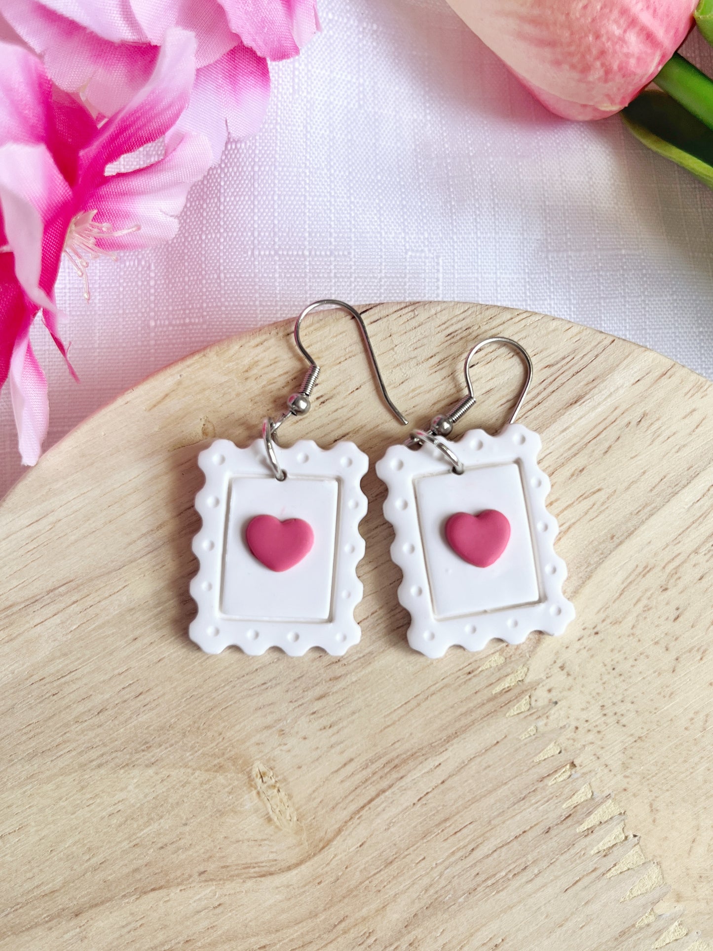 Heart Stamp Polymer Clay Earrings by silverstar.nz, shaped like white postage stamps with pink heart centers, dangle elegantly on stainless steel hooks on a wooden surface. Partial pink flowers in the background enhance the scenes charm.