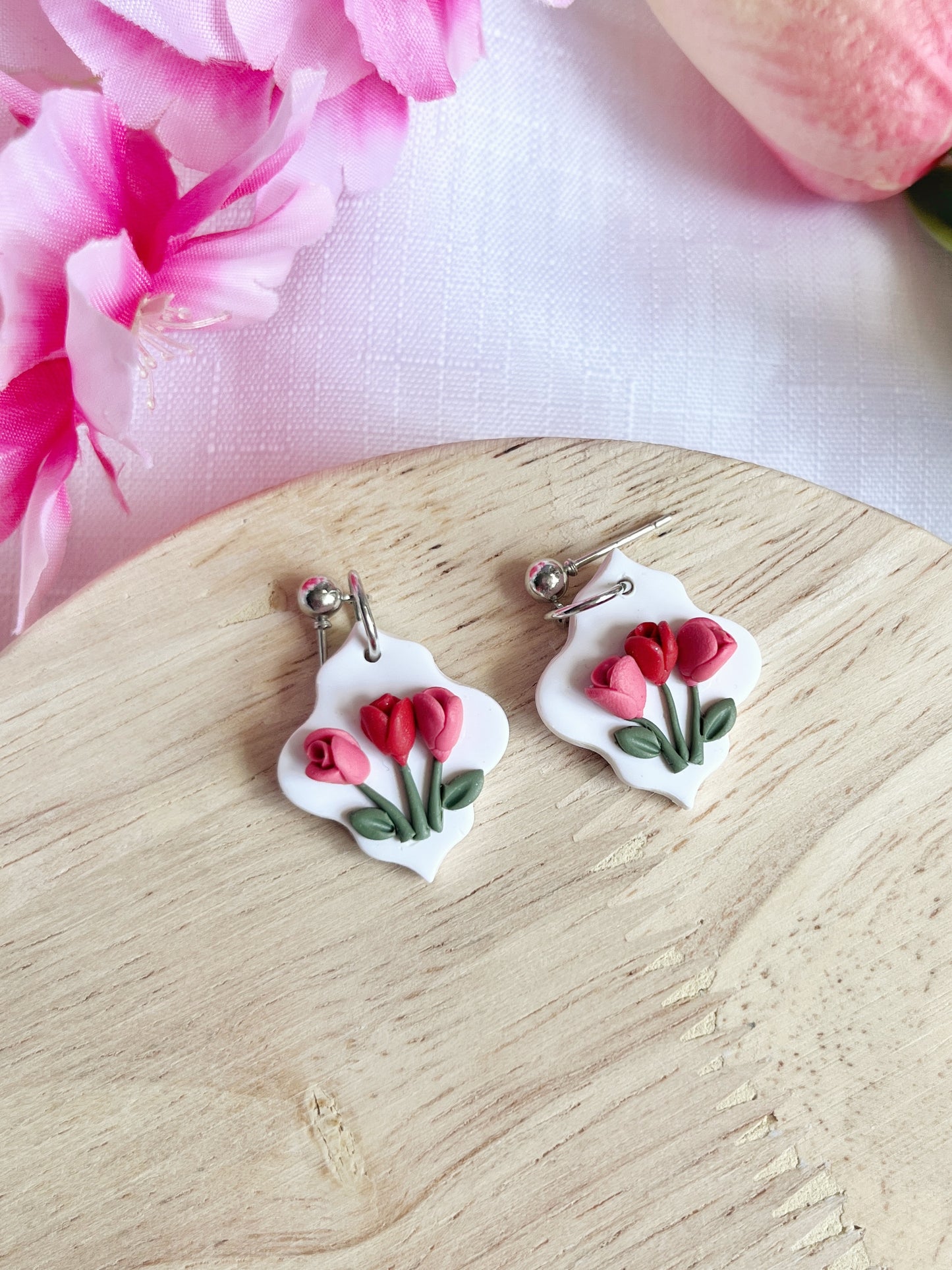 The Tulip Floral Polymer Clay Earrings by silverstar.nz, with delicate pink and red flowers, rest gracefully on a wooden surface. Their stainless-steel hooks guarantee durability, complemented by nearby pink flowers adding a soft, floral touch to the scene.