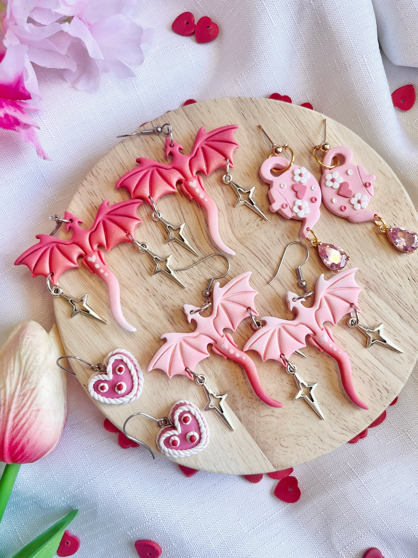 A wooden platter displays dragon-shaped earrings, heart cookies, and silverstar.nzs Love Tea Pot Polymer Clay Earrings with flower designs. Surrounding the platter are pink flowers, a tulip, and tiny red hearts scattered on light fabric.
