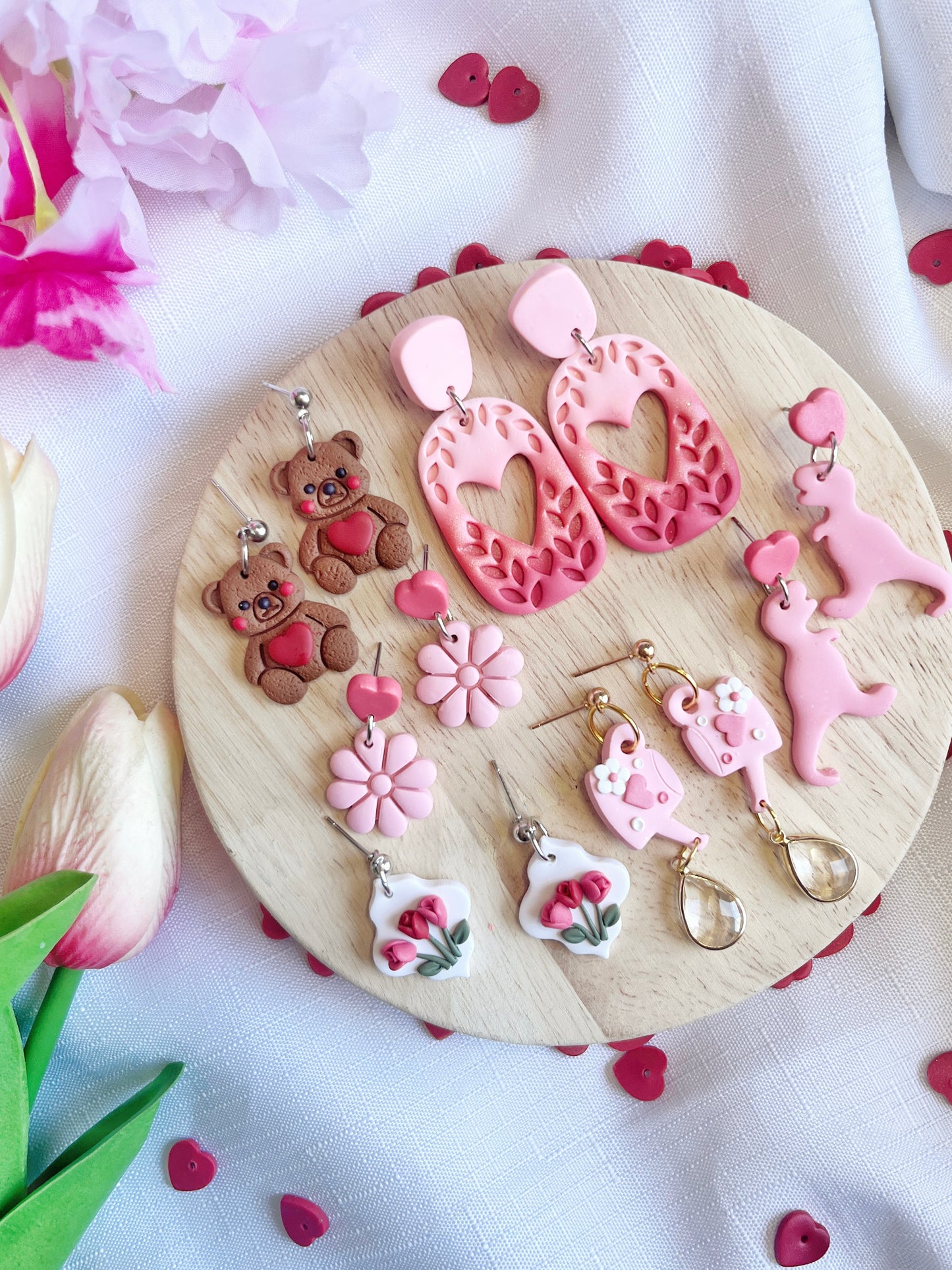 On a wooden platter, various handmade earrings are showcased, including silverstar.nzs Love Watering Can Polymer Clay Earrings. The display is accentuated with pink tulips, red heart confetti, and partial pink flowers on a white cloth.