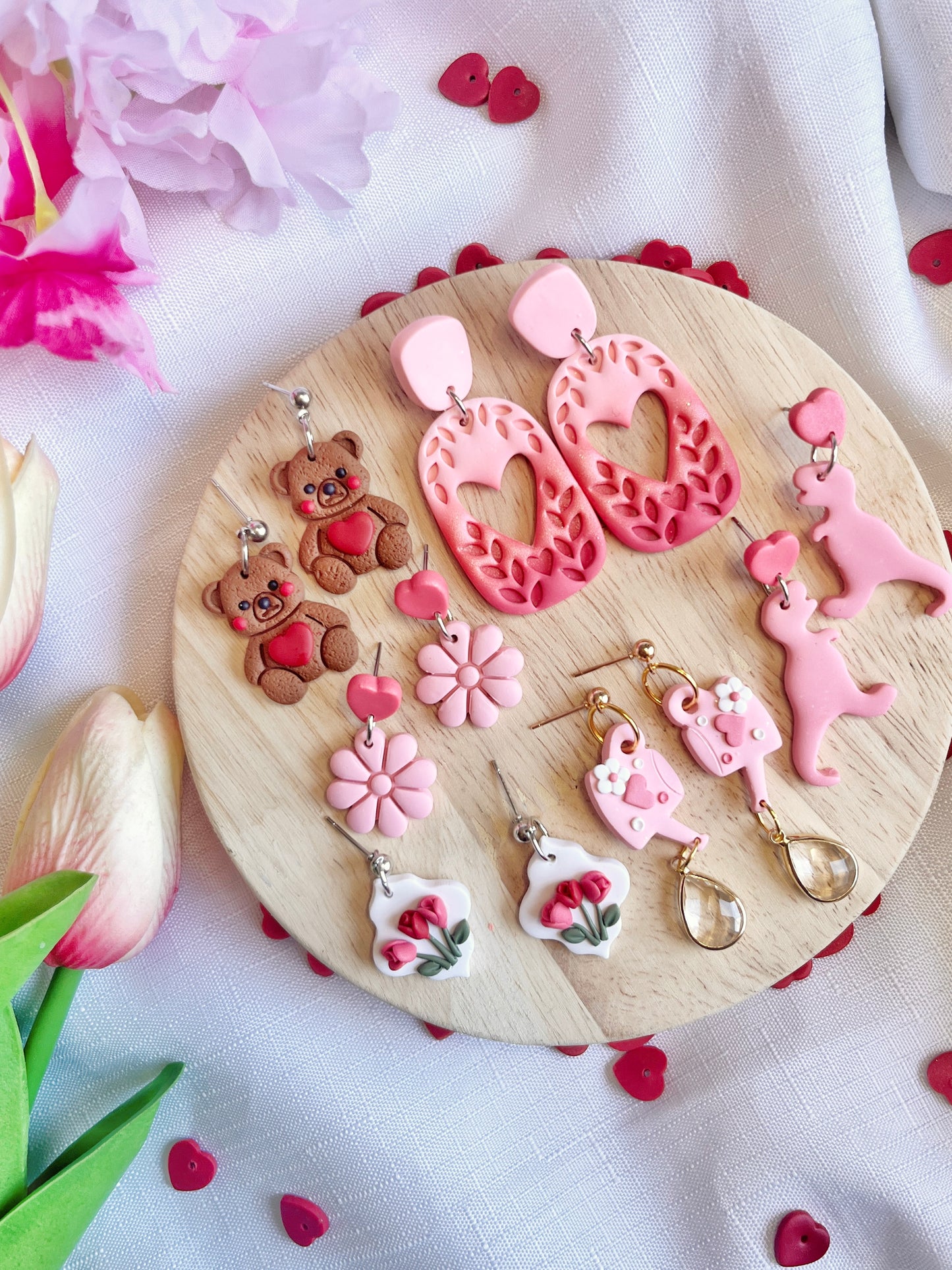 A wooden platter displays six pairs of silverstar.nzs Leafy Heart Dangle Polymer Clay Earrings on stainless steel hooks in fun designs: teddy bears, pink hearts, flowers, pink poodles, roses, and teardrops. A white cloth background is decorated with pink tulips and small red hearts.