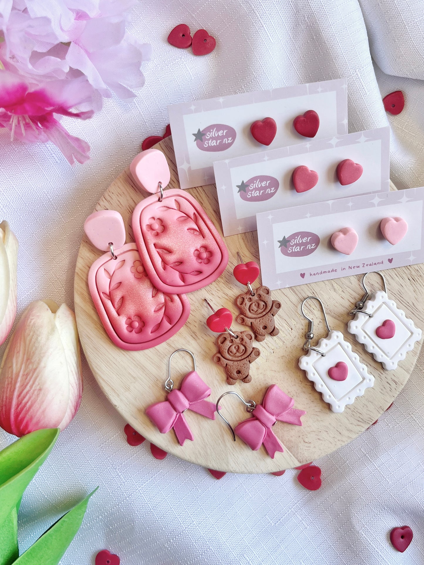Silverstar.nz displays their Valentines Heart Polymer Clay Stud Earrings on a wooden platter. The collection includes heart, pink bow, and floral rectangular earrings, along with teddy bear and frame designs. Pink flowers and green leaves enhance the funky setup.