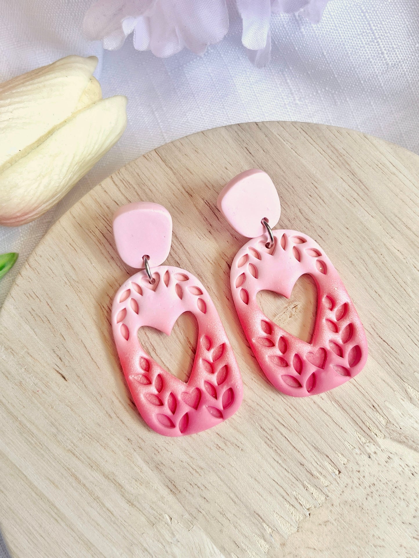 The Leafy Heart Dangle polymer clay earrings by silverstar.nz, featuring unique pink and red funky heart and leaf cutout designs, rest on a light wooden surface. They have durable stainless steel hooks with a partially visible white flower in the background.