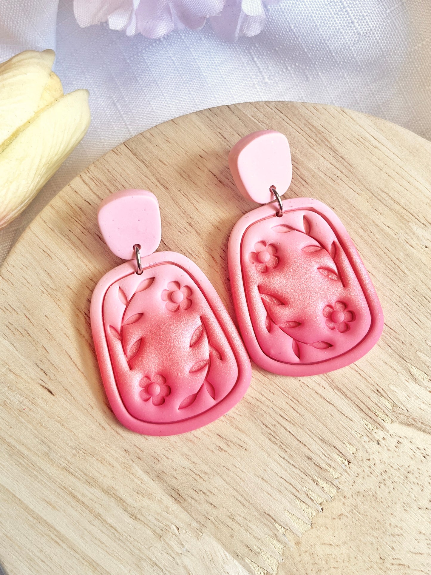 Statement Floral Polymer Clay Earrings from silverstar.nz feature a unique design with pink floral and leaf patterns on a rectangular shape with rounded edges, set against a wooden surface. These handmade earrings showcase darker pink flower and leaf designs paired with a lighter pink top piece.